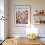 a kitchen scene with a framed poster of a colorful flower market in Omaha, Nebraska, hanging on the wall above a wooden counter. The poster is surrounded by various kitchen items, such as jars, a bowl of lemons, and a plant.