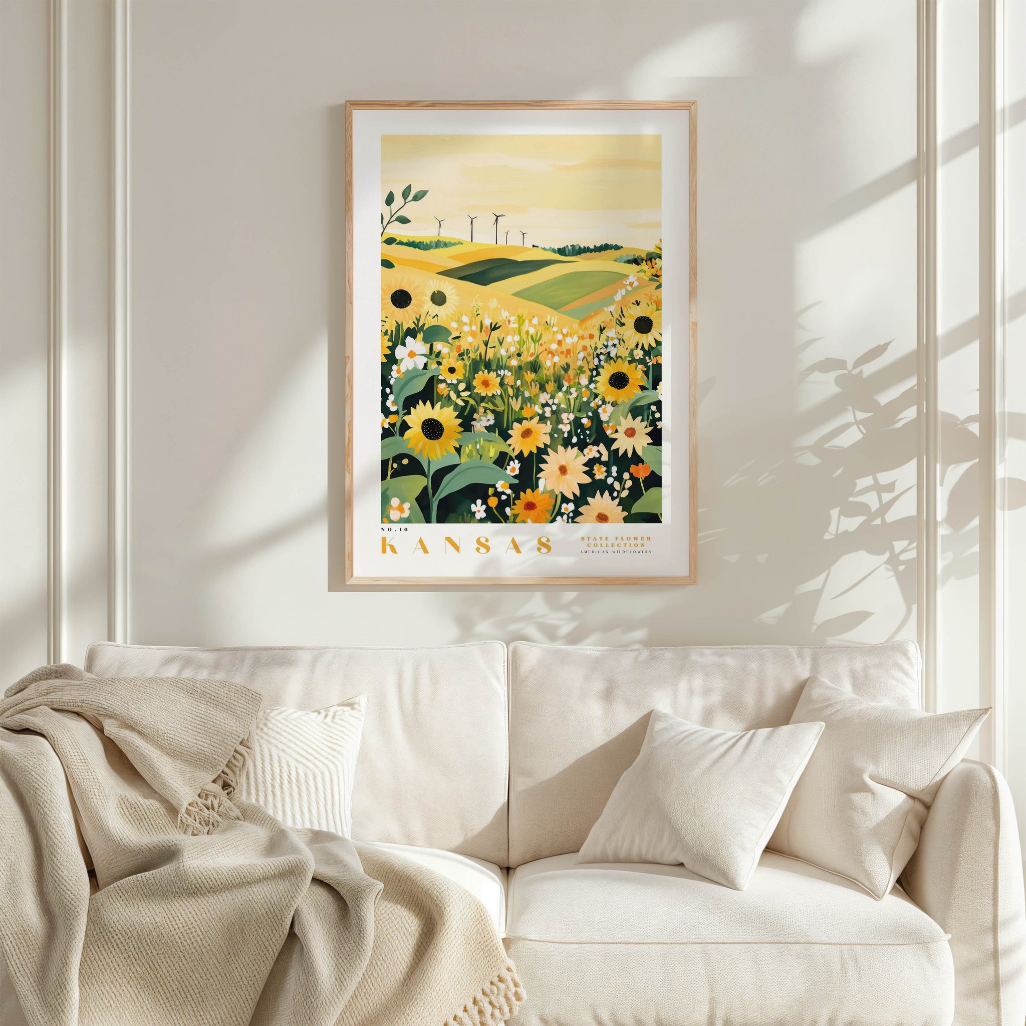 a framed poster of a field with sunflowers hanging on a wall above a white couch with a blanket draped over it.