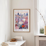a framed poster of a cityscape with a colorful floral design, hanging on a wall in a room with a window and a small table with books and a vase.