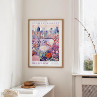 a framed poster of a cityscape with a colorful floral design, hanging on a wall in a room with a window and a small table with books and a vase.