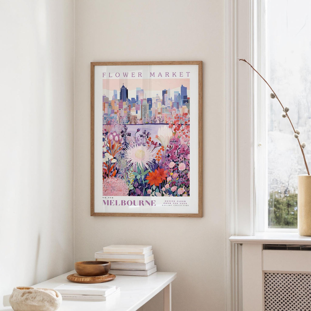 a framed poster of a cityscape with a colorful floral design, hanging on a wall in a room with a window and a small table with books and a vase.