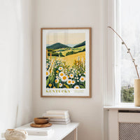 a framed poster of a field with flowers hanging on a wall, with a white table and a vase of flowers in the foreground.