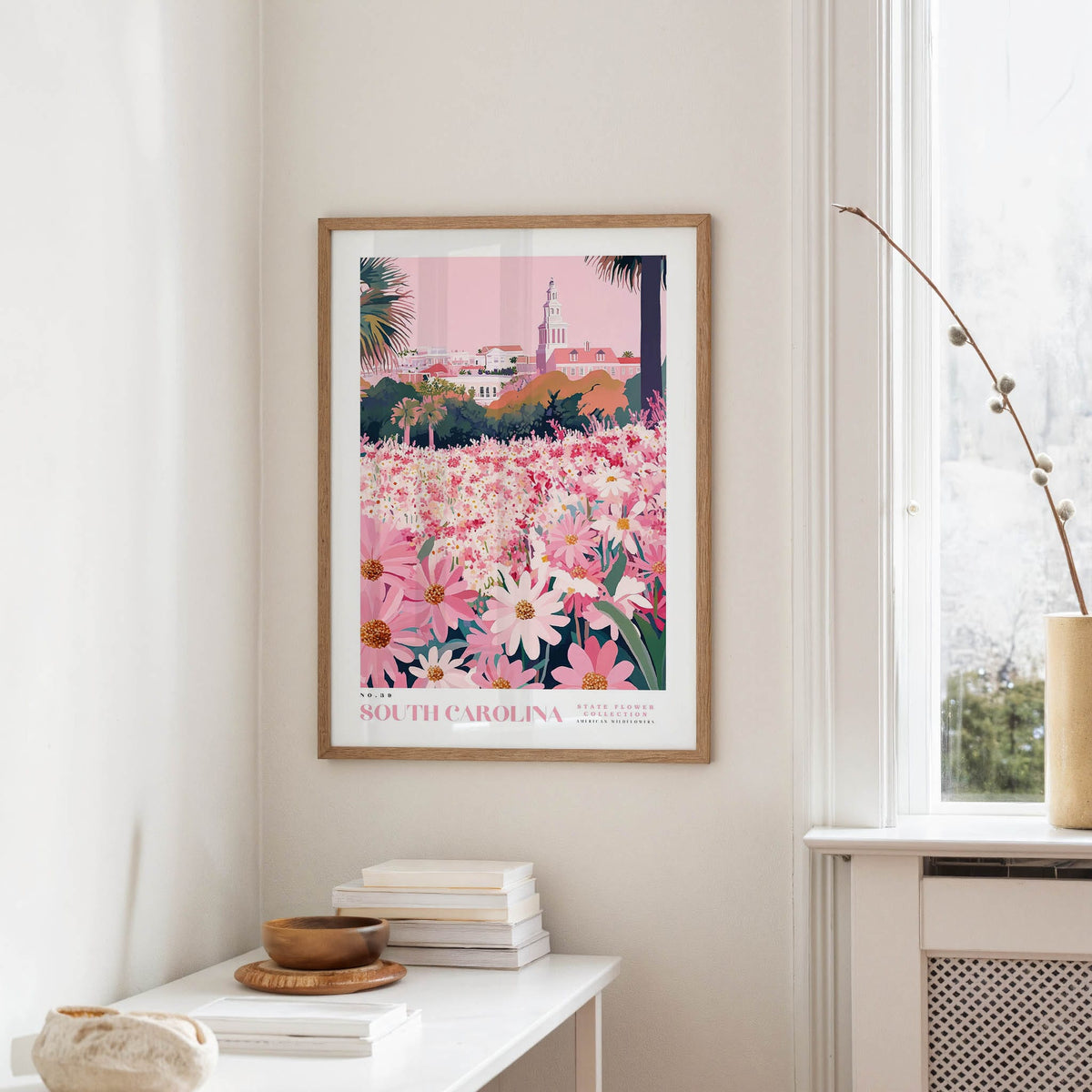 a framed poster of a colorful floral scene hanging on a wall, with a white table and a vase of flowers in the foreground.