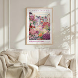 a framed poster of a colorful garden scene hanging on a wall above a white couch with a blanket draped over it.