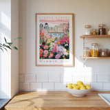 a framed poster of a colorful garden scene with flowers and buildings, hanging on a wall above a wooden table with a bowl of lemons.