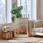 a living room with a large framed poster of a colorful floral scene hanging on the wall, a stack of books on a wooden shelf, a white sofa, and a large window with a view of buildings outside.
