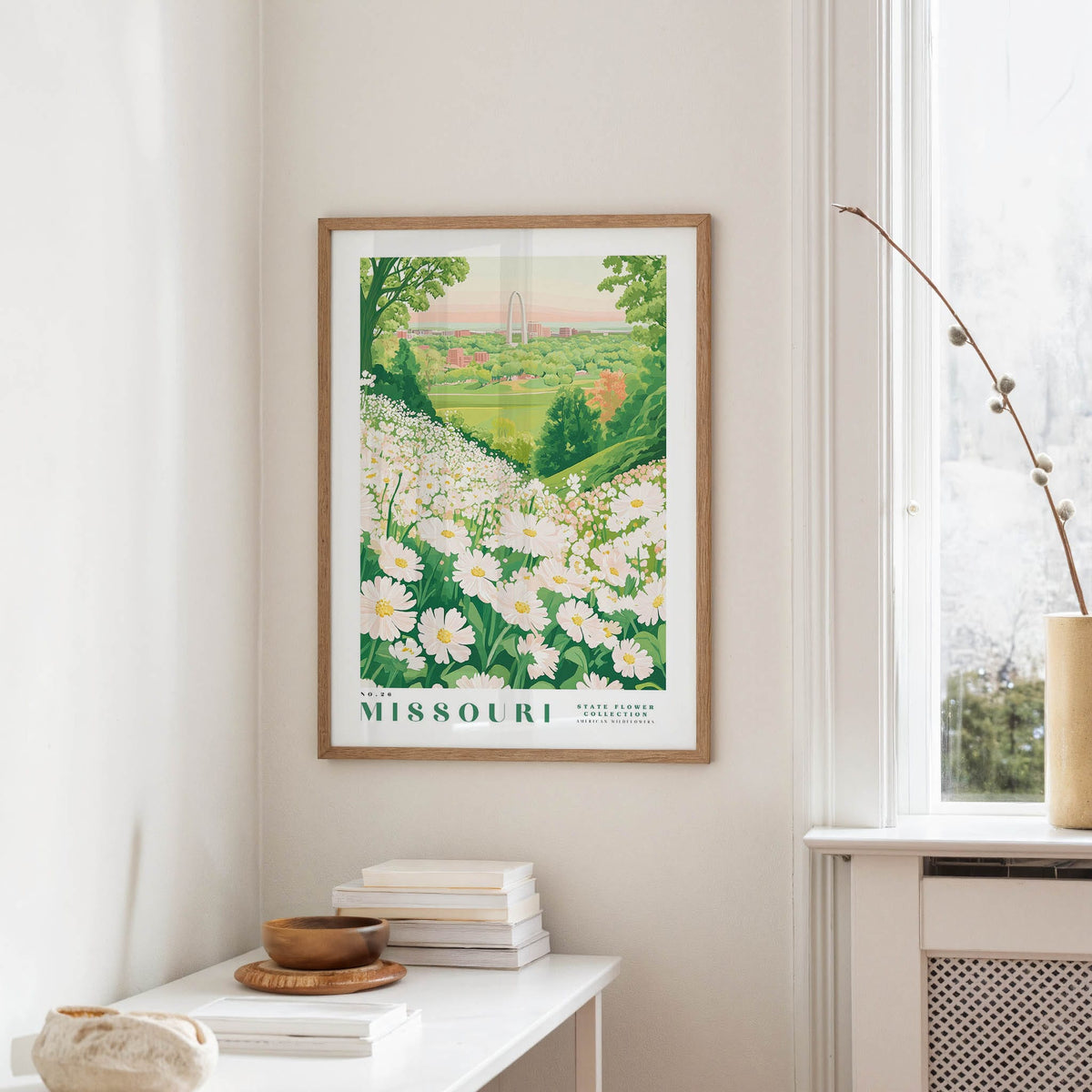 a framed poster of a field of white flowers, with the text "Missouri" prominently displayed. The poster is hanging on a wall, and there is a small table with a stack of books and a vase nearby.