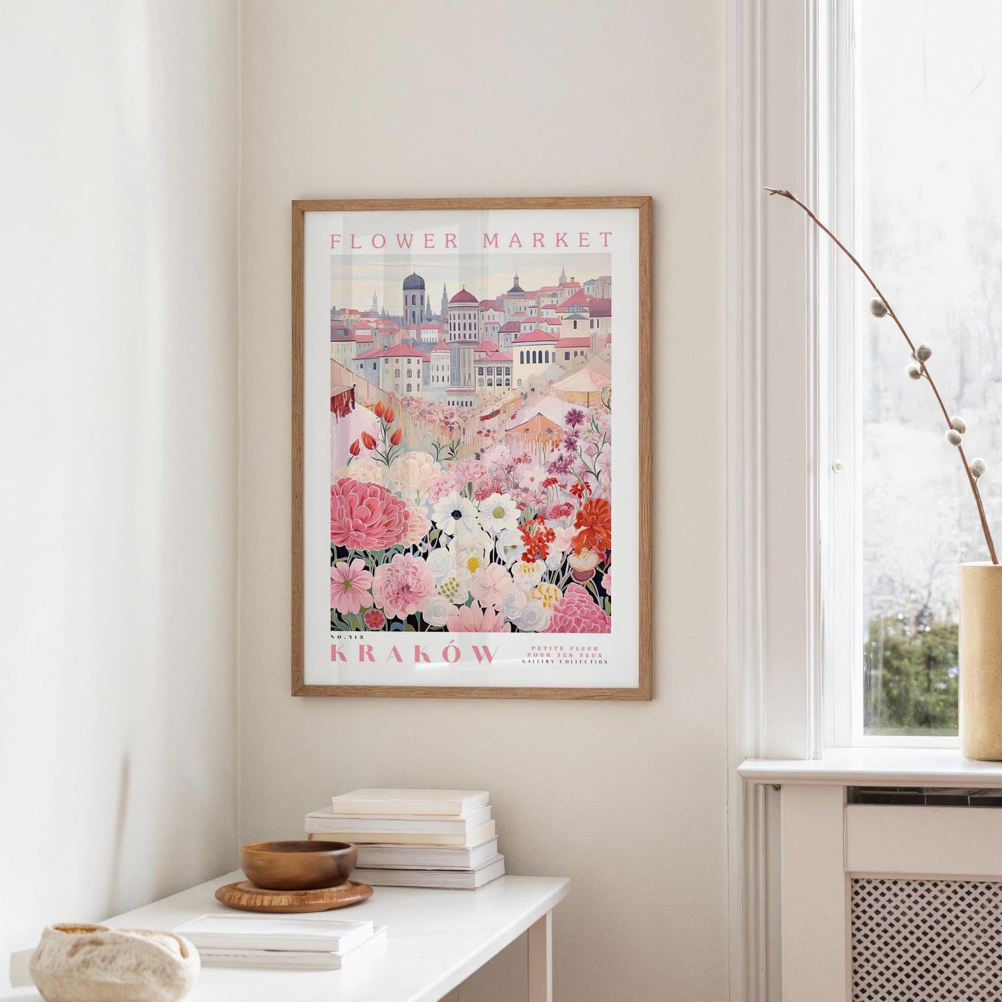 a framed poster of a cityscape with a flower market in the background, hanging on a wall in a room with a window and a table with books and a bowl on it.