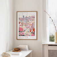 a framed poster of a cityscape with a flower market in the background, hanging on a wall in a room with a window and a table with books and a bowl on it.