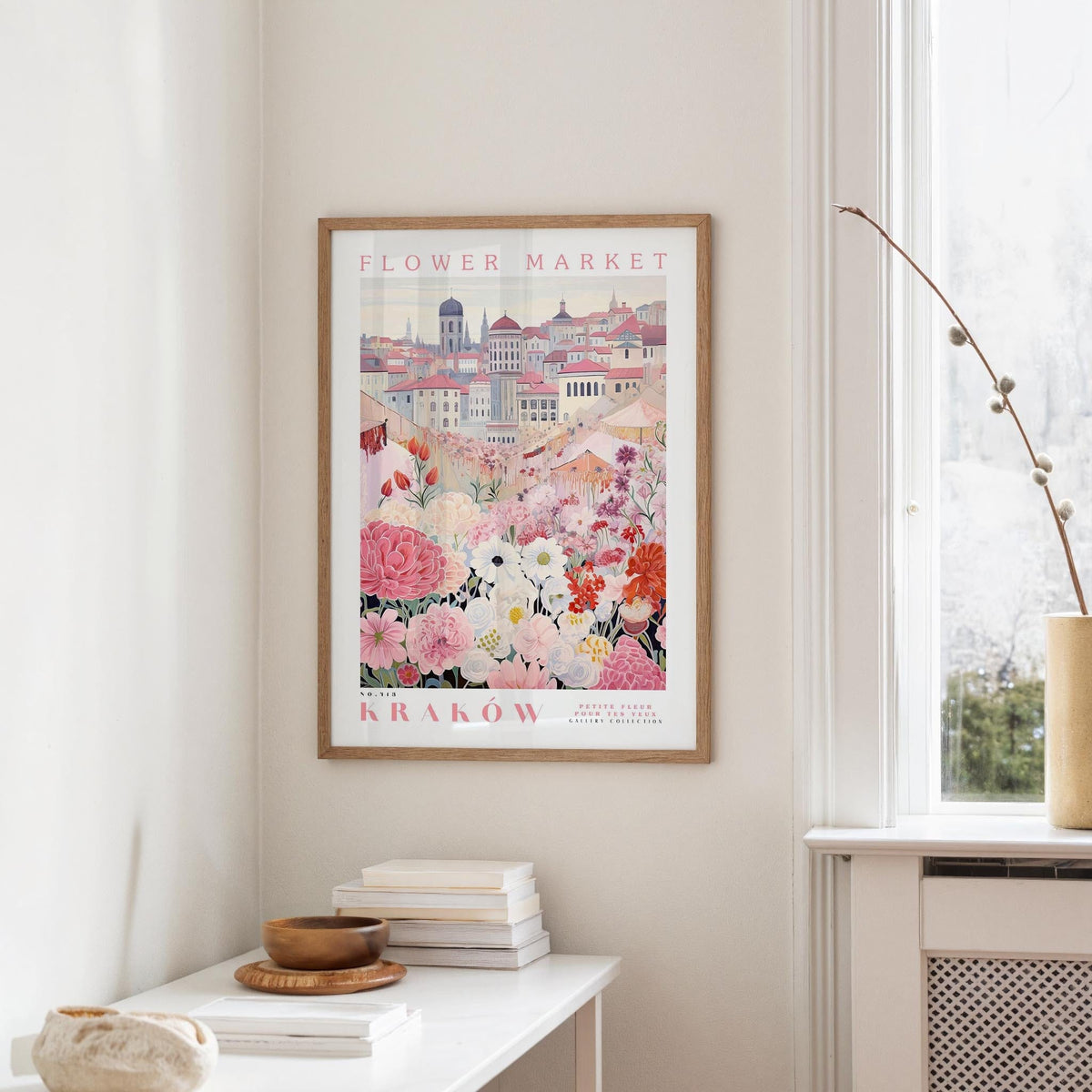a framed poster of a cityscape with a flower market in the background, hanging on a wall in a room with a window and a table with books and a bowl on it.