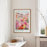 a framed poster of a colorful floral scene hanging on a wall, with a white table and a vase of flowers in the foreground.