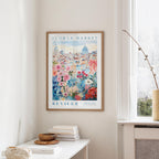 a framed poster of a cityscape with a flower market in the background, hanging on a wall in a room with a window and a table with books and a vase.