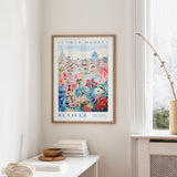 a framed poster of a cityscape with a flower market in the background, hanging on a wall in a room with a window and a table with books and a vase.