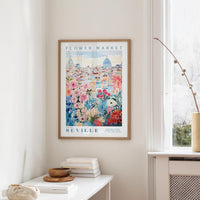 a framed poster of a cityscape with a flower market in the background, hanging on a wall in a room with a window and a table with books and a vase.