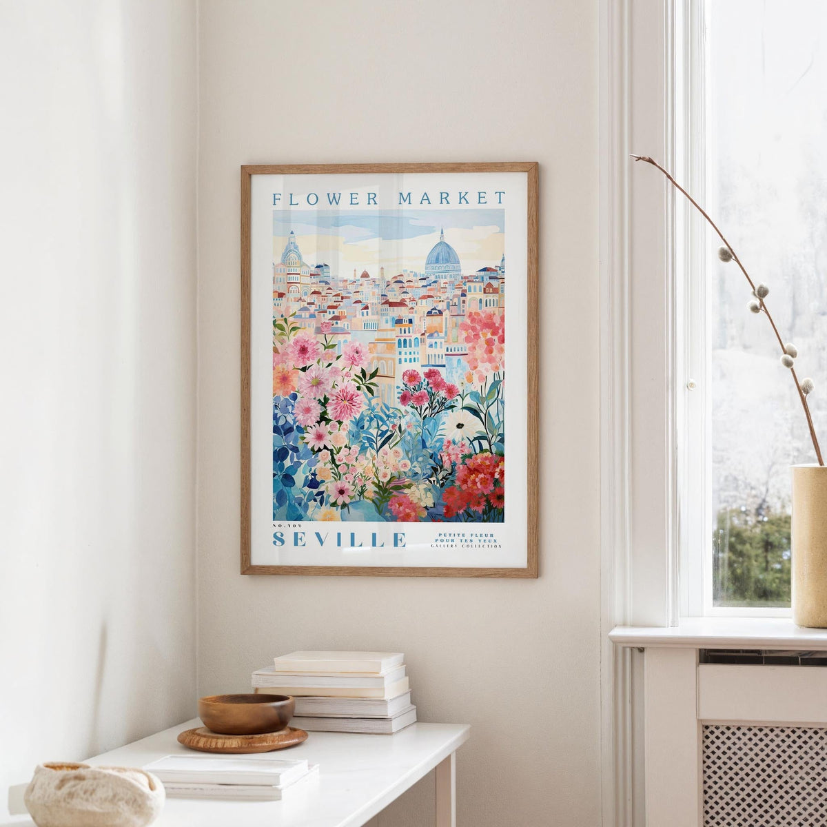 a framed poster of a cityscape with a flower market in the background, hanging on a wall in a room with a window and a table with books and a vase.