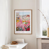 a framed poster of a colorful floral scene hanging on a wall, with a white table and a vase of flowers in the foreground.