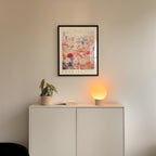 a white cabinet with a potted plant and a lamp on top, set against a wall with a framed poster of a cityscape above it.