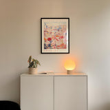 a white cabinet with a potted plant and a lamp on top, set against a wall with a framed poster of a cityscape above it.