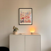 a white cabinet with a potted plant and a lamp on top, set against a wall with a framed poster of a cityscape above it.