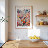 a kitchen counter with a bowl of lemons, a framed poster of a flower market, and a shelf with jars and other kitchen items.