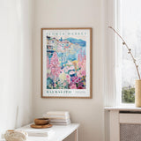 a framed poster of a colorful floral scene hanging on a wall, with a white table and a vase of flowers in the foreground.