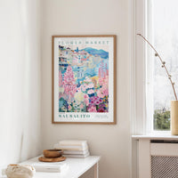 a framed poster of a colorful floral scene hanging on a wall, with a white table and a vase of flowers in the foreground.