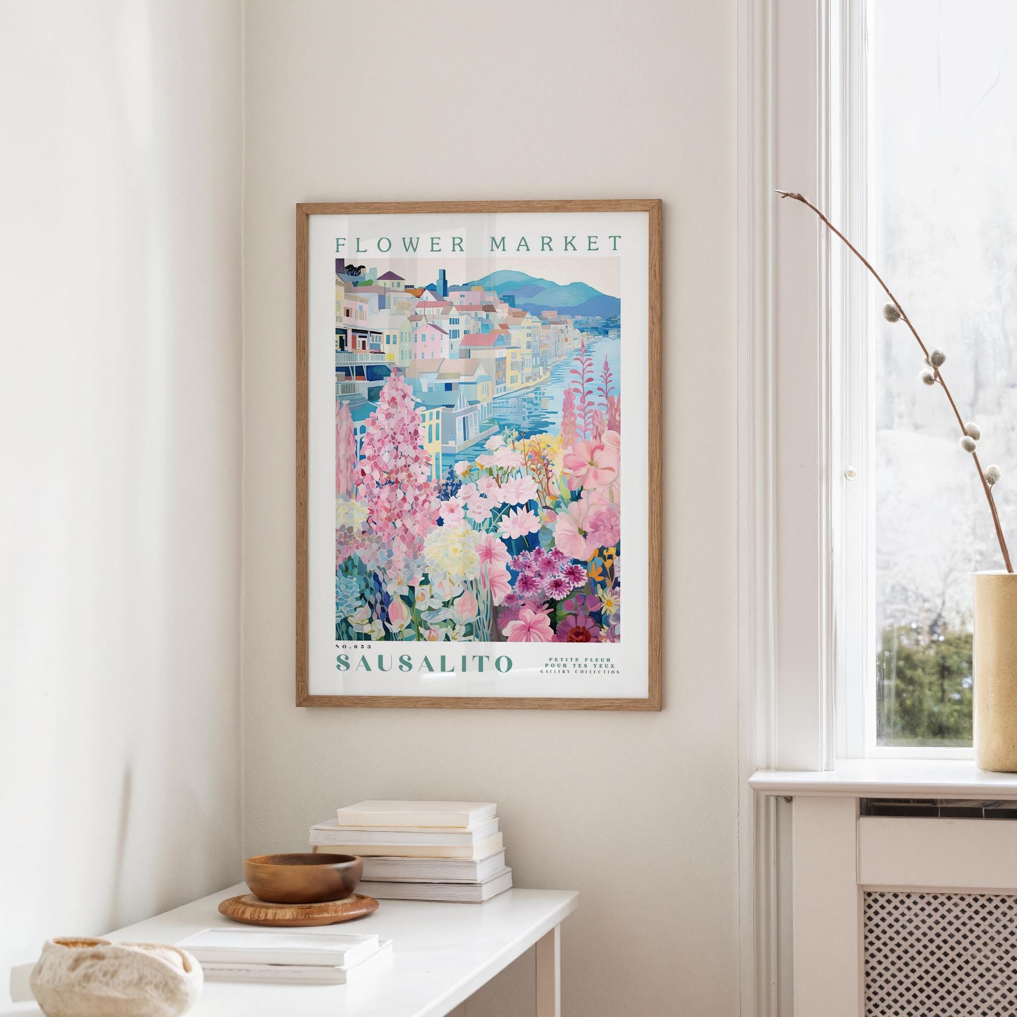 a framed poster of a colorful floral scene hanging on a wall, with a white table and a vase of flowers in the foreground.