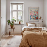 a cozy bedroom with a large bed, a wooden nightstand, and a window with a plant on the sill. The room has a warm, inviting atmosphere with a neutral color palette and natural elements like plants and a rug.
