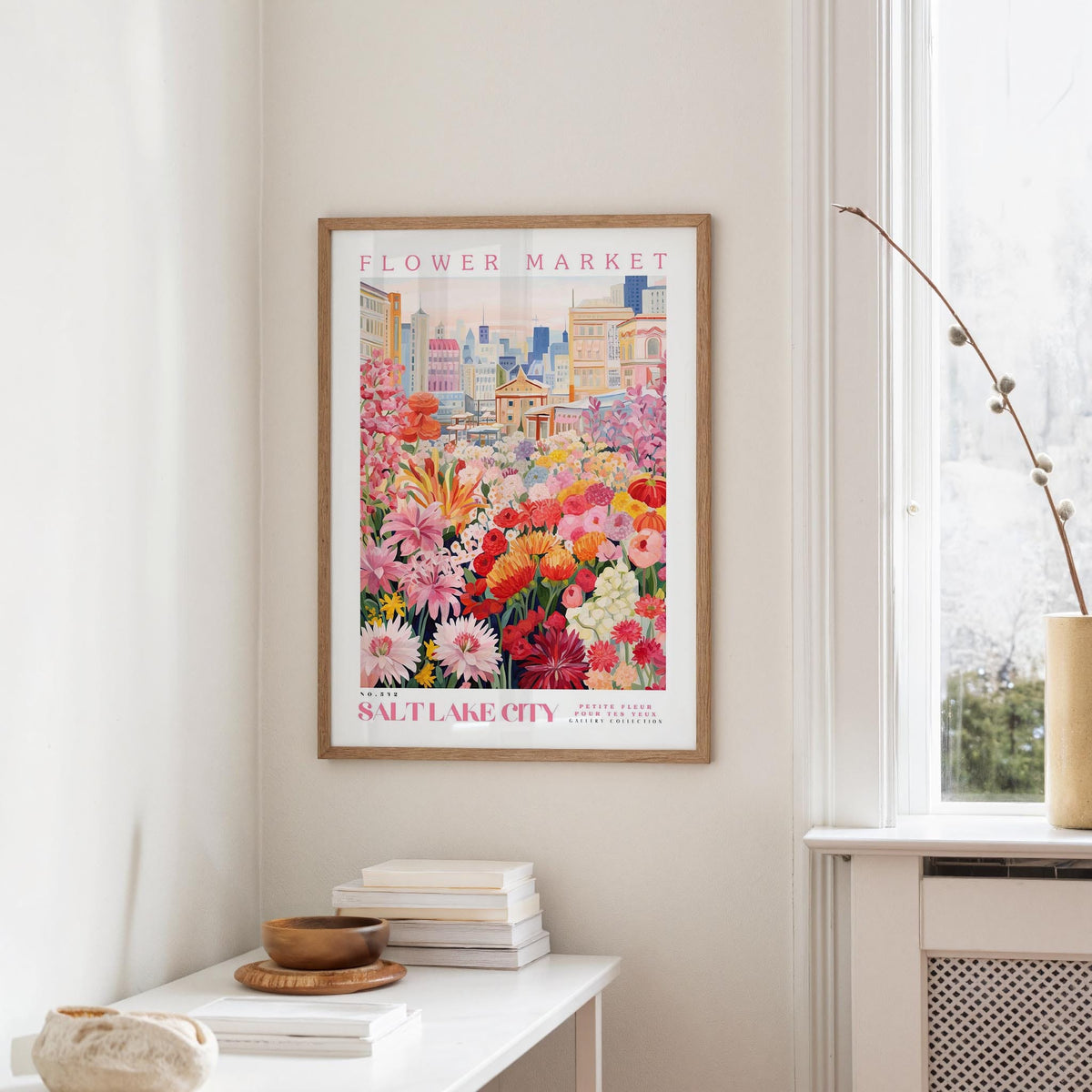 a framed poster of a colorful cityscape with flowers hanging on a wall, next to a window and a small table with books and a bowl.