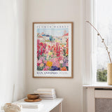 a framed poster of a colorful garden scene hanging on a wall, with a white table and a vase of flowers in the foreground.