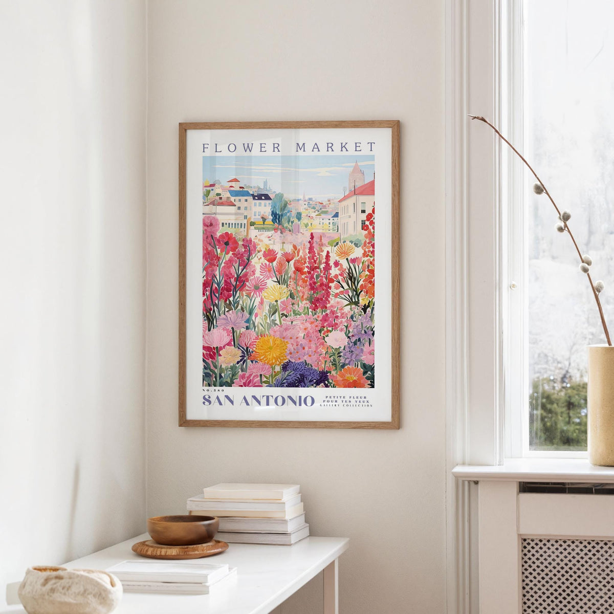 a framed poster of a colorful garden scene hanging on a wall, with a white table and a vase of flowers in the foreground.