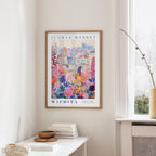a framed poster of a colorful cityscape with flowers hanging on a wall in a room with a white table and a window.
