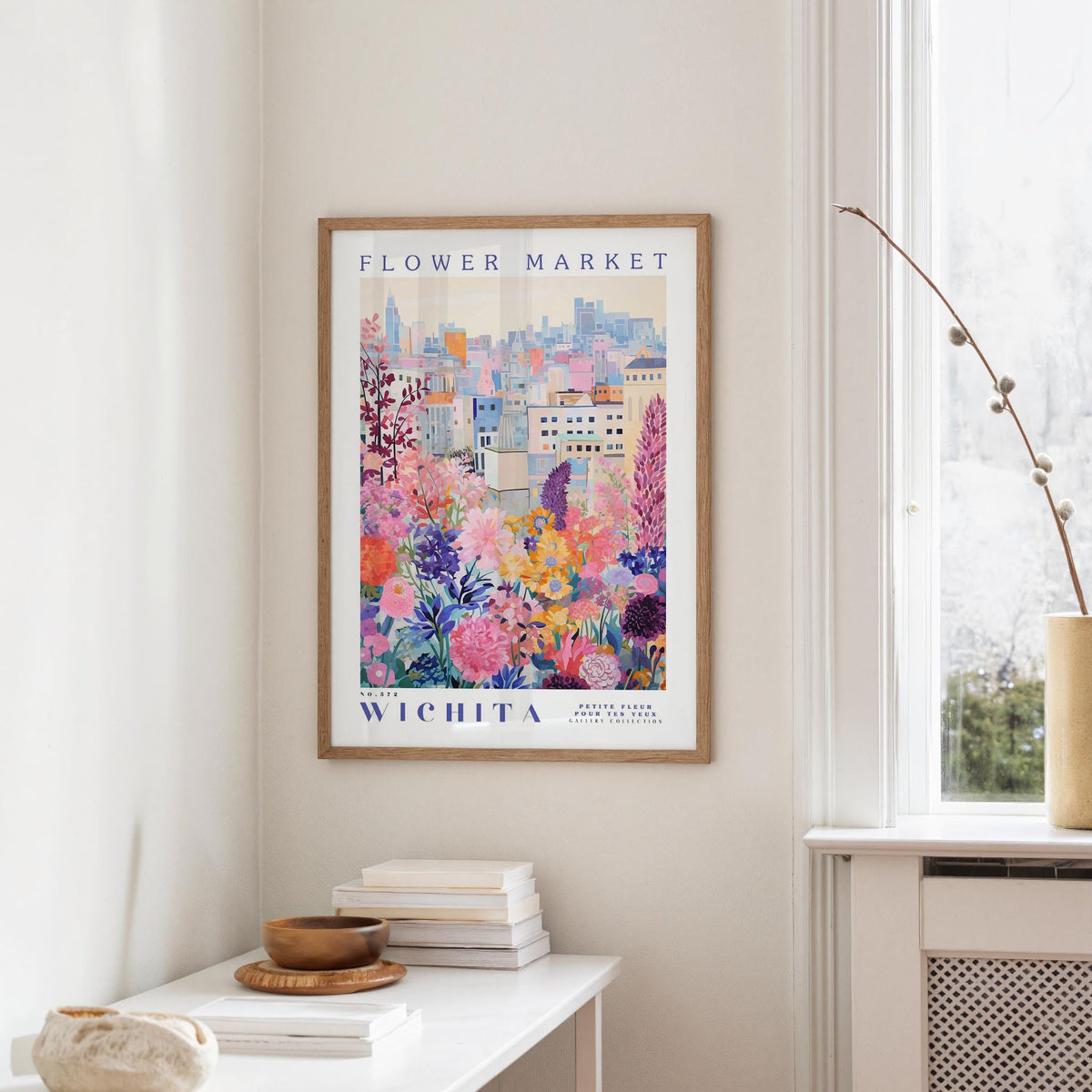 a framed poster of a colorful cityscape with flowers hanging on a wall in a room with a white table and a window.