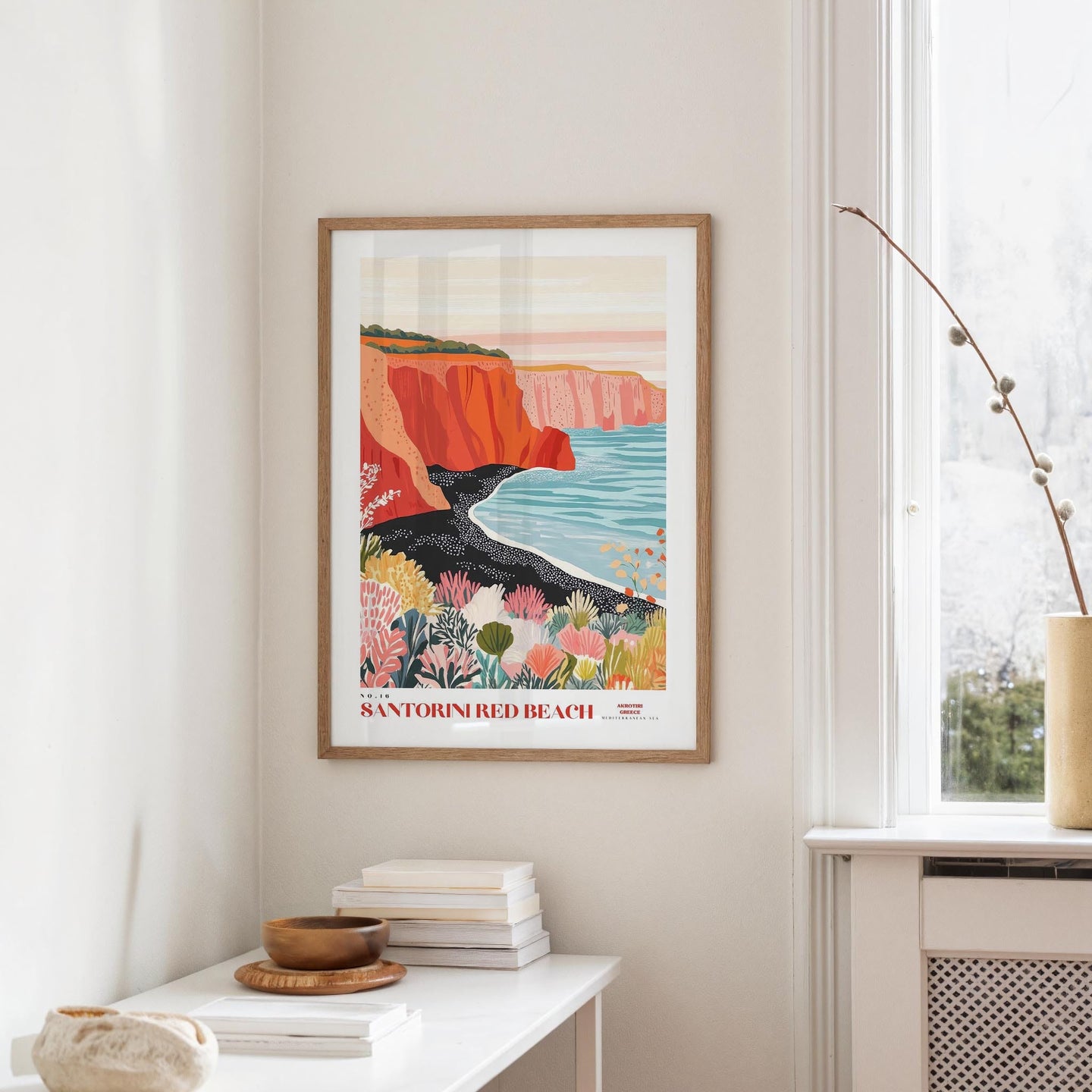 a framed poster of Santorini's red beach, hanging on a wall in a room with a white table and a window.