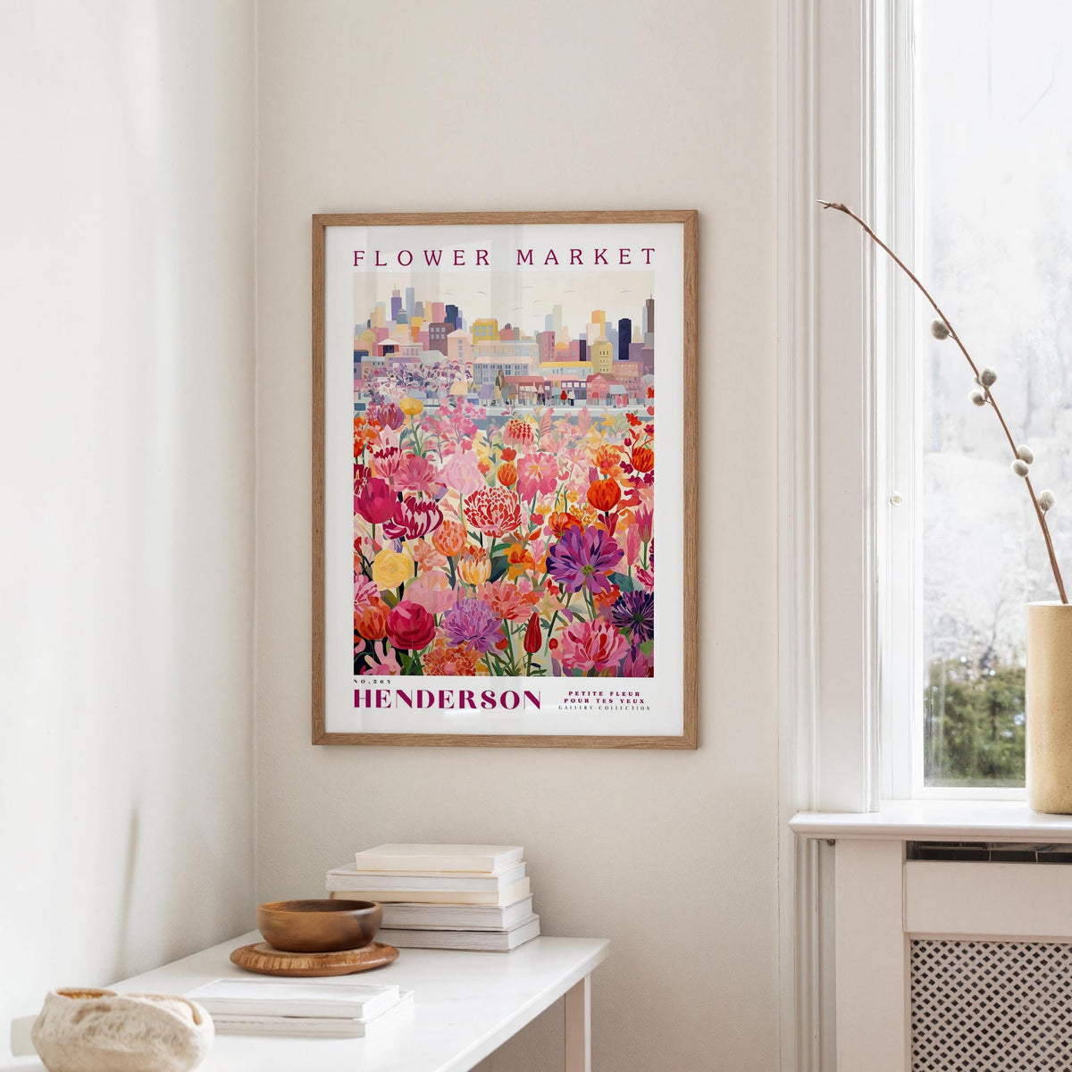 a framed poster of a colorful flower market scene hanging on a wall, with a white table and a vase of flowers in the foreground.