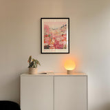 a white cabinet with a potted plant and a lamp on top, set against a wall with a framed poster of a tropical island.