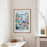 a framed poster of a colorful floral scene hanging on a wall, with a white table and a vase of flowers in the foreground.