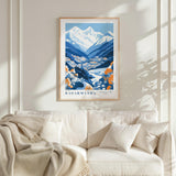 a framed poster of a mountainous landscape with a village in the valley, hanging on a wall above a white couch with a blanket draped over it.