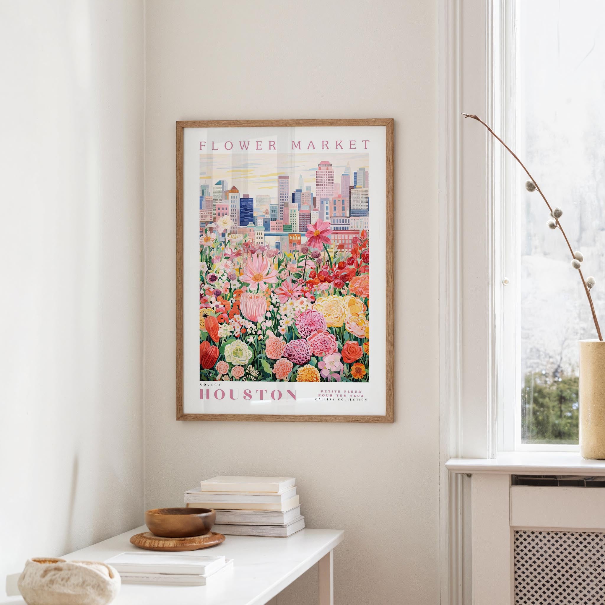 a framed poster of a colorful cityscape with a flower market in the background, hanging on a wall in a room with a window and a small table with books and a vase.