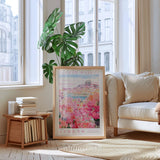 a living room with a large framed poster of a flower market in Athens hanging on the wall, a stack of books on a table, a white sofa, and a large window with a view of the city outside.