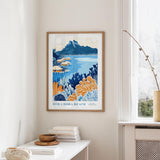 a framed poster of a tropical beach scene, with a mountain in the background and colorful flowers in the foreground. The poster is hanging on a wall, and there is a table with books and a vase nearby.