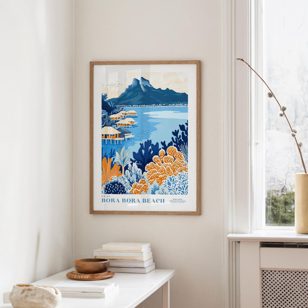 a framed poster of a tropical beach scene, with a mountain in the background and colorful flowers in the foreground. The poster is hanging on a wall, and there is a table with books and a vase nearby.