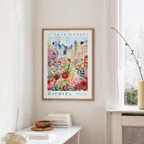 a framed poster of a colorful cityscape with flowers, hanging on a wall in a room with a white table and a window.