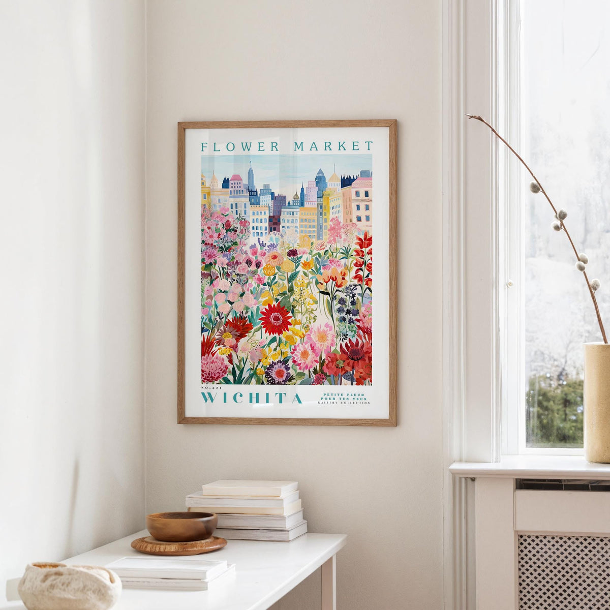 a framed poster of a colorful cityscape with flowers, hanging on a wall in a room with a white table and a window.