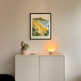 a framed poster of a beach scene hanging on a wall above a white cabinet with a plant and a lamp on top.
