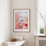 a framed poster of a colorful floral scene hanging on a wall, with a window and a small table with books and a vase nearby.