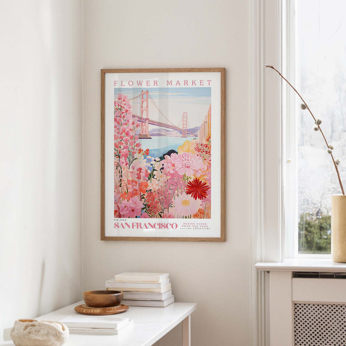 a framed poster of a colorful floral scene hanging on a wall, with a window and a small table with books and a vase nearby.