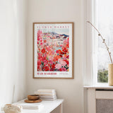 a framed poster of a colorful floral scene hanging on a wall, with a white table and a vase of flowers in the foreground.