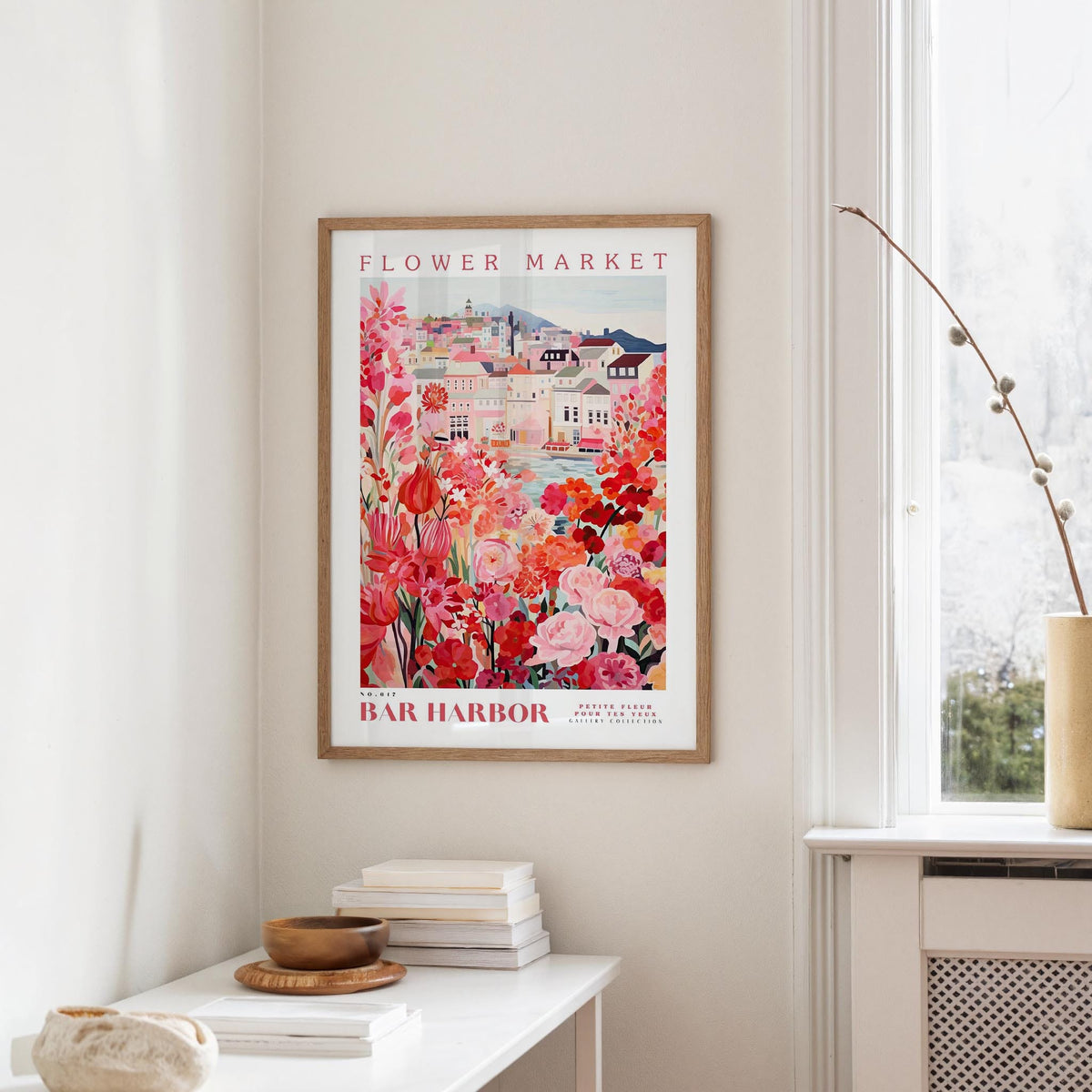 a framed poster of a colorful floral scene hanging on a wall, with a white table and a vase of flowers in the foreground.