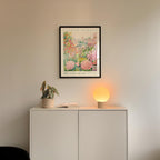 a white cabinet with a potted plant and a lamp on top, set against a wall with a framed poster of a garden scene hanging above it.